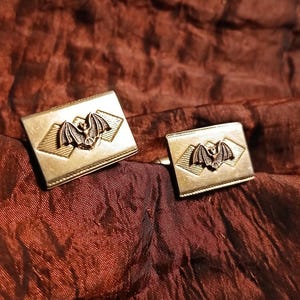 May include: A pair of gold-toned cufflinks with a bat design. Each cufflink features a rectangular shape with a central bat emblem, set within a diamond-shaped border. The cufflinks are displayed on a textured, reddish-brown fabric.