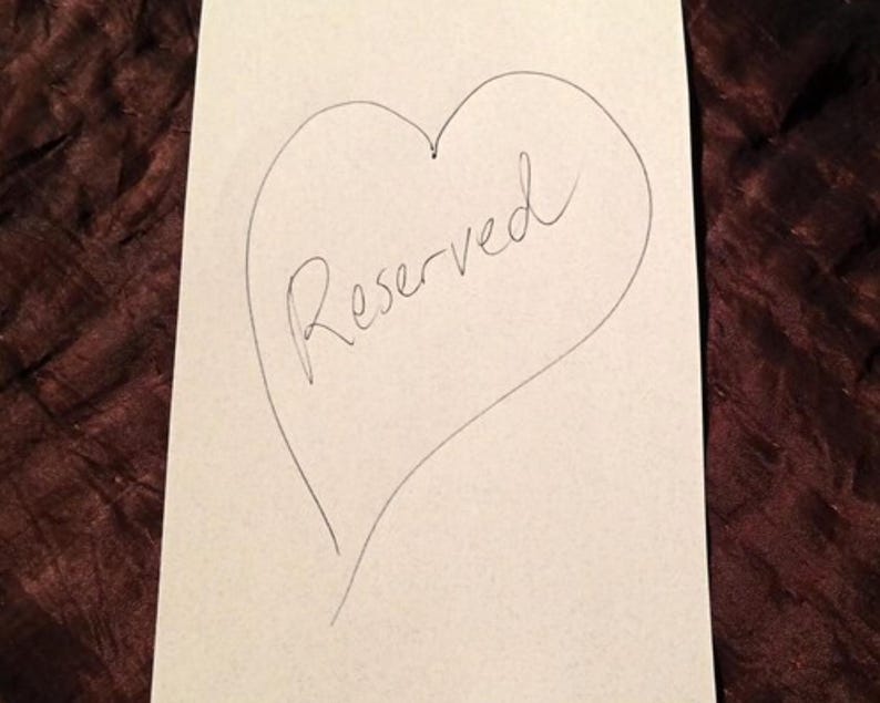 Puede incluir: Una tarjeta blanca con un coraz&oacute;n dibujado a mano y la palabra "Reserved" escrita en cursiva en su interior. La tarjeta est&aacute; sobre un fondo oscuro y texturizado.