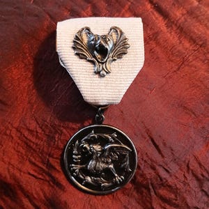 Peut inclure: Médaille vintage avec une finition argentée. La médaille présente un motif détaillé d'une créature mythique sur un pendentif circulaire, suspendu à un ruban blanc. Le ruban est attaché à un emblème décoratif argenté.