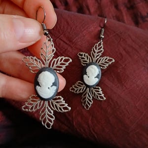 May include: A pair of silver-toned earrings featuring oval black cameos with white profile portraits. The cameos are set within a filigree leaf design. The earrings are held up by a silver fish hook.