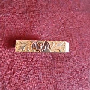 Puede incluir: Un pasador de corbata de metal dorado con un diseño de murciélago en el centro. El murciélago es de color cobre y el pasador tiene un patrón floral grabado. El fondo es una tela de color rojo oscuro.