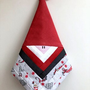 May include: A red and white Christmas gnome ornament with a white beard and a red hat. The gnome is made of fabric and has a white and gray patterned background with small gnomes.