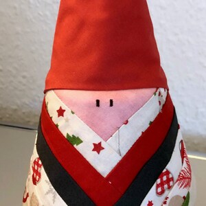 May include: A red and white Christmas gnome with a red hat and a pink face. The gnome is made of fabric and has a patchwork design with stars, pine trees, and other Christmas motifs.