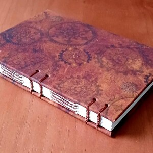 May include: A brown leather-bound journal with a steampunk design featuring a pattern of gears and cogs. The journal is closed and lying on a wooden surface.