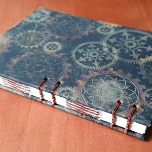 May include: A dark blue journal with a steampunk design featuring intricate gears and patterns. The cover is bound with copper wire and has a white spine.