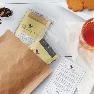 May include: Overhead shot of a tea set with a brown paper bag, tea packets, a glass of tea, and biscuits. The tea packets are labelled "Plum Deluxe" and "Black Tea". A card reads "Tea of the Month Cinnamon Nourish".