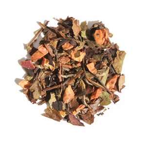 May include: A close-up of a blend of white tea leaves, dried apple pieces, and other botanicals. The tea leaves are a light green colour, and the apple pieces are a light brown colour.
