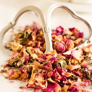 May include: A heart-shaped tea infuser made of silver, filled with dried rose petals. The petals are a mix of pink, red, and yellow, spilling onto a white surface. The infuser is open, suggesting use for loose leaf tea.