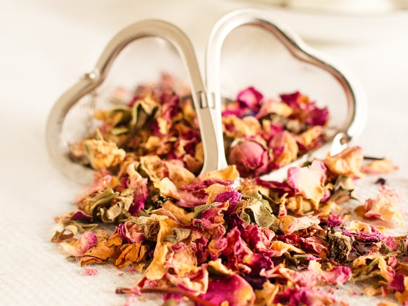 May include: A heart-shaped tea infuser made of silver, filled with dried rose petals. The petals are a mix of pink, red, and yellow, spilling onto a white surface. The infuser is open, suggesting use for loose leaf tea.