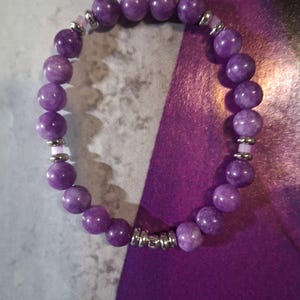 May include: A beaded bracelet featuring round, purple beads. The bracelet has silver-colored spacers and accents. The beads are a deep, solid purple color. The bracelet is displayed on a purple and gray background.
