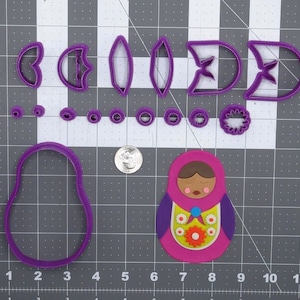 May include: Purple plastic cookie cutters in the shape of a nesting doll, flowers, and leaves. A pink fondant nesting doll is also shown.