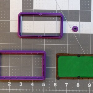 Op de afbeelding: Drie paarse plastic koekjesvormen in de vorm van een biljarttafel, een biljartbal en een rechthoek. De biljarttafelvorm heeft een bruin vilt oppervlak en zwarte stippen voor de zakken.