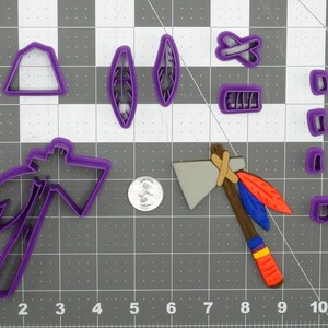 May include: A set of purple plastic cookie cutters in the shape of a tomahawk, feathers, a teepee, and a cross. The cutters are on a gray gridded surface.