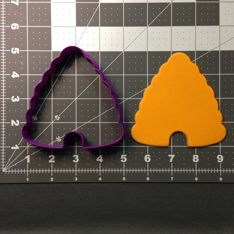 May include: Purple plastic cookie cutter shaped like a beehive with a yellow plastic beehive next to it on a black and white grid.