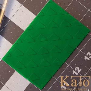 Può includere: Tappetino di embossing in plastica verde con un motivo ripetitivo di piccoli triangoli. Il tappetino è su una superficie a griglia grigia e bianca.