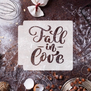 May include: White stencil with the text "Fall in love" written in a cursive font. The stencil is on a brown surface with flour scattered around it.