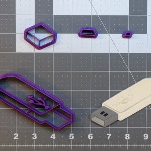 Puede incluir: Cortadores de galletas de plástico morado con forma de unidad USB, cubo y dos rectángulos. También se muestra un cortador de galletas de unidad USB de plástico blanco.