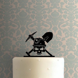 Può includere: Silhouette nera di uno scheletro che indossa un casco da minatore con una piccozza e una pala incrociate dietro di lui. Il topper è adatto per una torta.