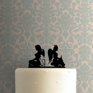 May include: A black silhouette of an angel and a devil cake topper. The angel is on the right and the devil is on the left. The angel has a halo and wings. The devil has horns and a tail.