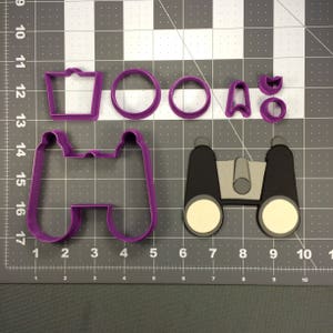 May include: Purple plastic cookie cutters in the shape of binoculars, a hat, and two circles. The cutters are on a gray grid background.