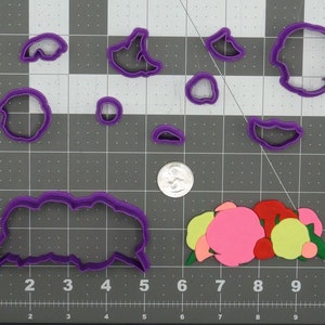 Puede incluir: Un conjunto de cortadores de galletas de plástico morado con forma de flor en varios tamaños y formas, junto con un arreglo floral de papel colorido. Los cortadores están dispuestos sobre una superficie de cuadrícula gris, con una moneda.