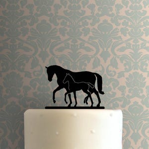 Puede incluir: Adorno para tarta con silueta negra que representa un caballo y un potro caminando juntos. El adorno para tarta está colocado sobre una tarta blanca. El fondo presenta un estampado floral en azul claro y beige. Un elemento decorativo.