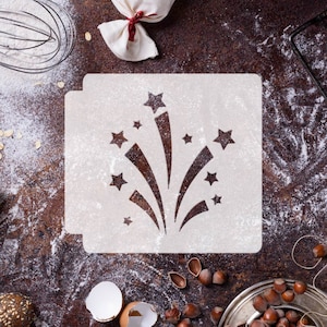 Può includere: Uno stencil di plastica bianca con un disegno di fuochi d'artificio. Lo stencil è su una superficie marrone con farina sparsa intorno.