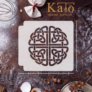 Könnte beinhalten: Eine weiße Kunststoffschablone mit einem keltischen Knotendesign. Die Schablone ist 12,7 cm breit und 12,7 cm hoch. Sie liegt auf einer braunen Oberfläche mit einem Lineal und anderen Backutensilien.