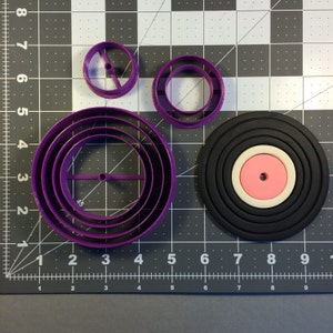 May include: A set of purple plastic cookie cutters in the shape of a record, with a smaller circle in the center. The cutters are on a gray grid background.