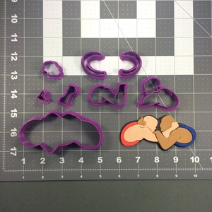 May include: Purple plastic cookie cutters in various shapes, including a heart, a circle, a figure eight, a pair of hands clasped together, and a cloud. The cutters are on a gray gridded surface.