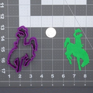 May include: Two cookie cutters, one purple and one green, shaped like a cowboy riding a bucking bronco. The cutters are on a gray gridded surface.
