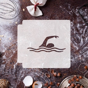 May include: A white stencil featuring a silhouette of a swimmer in the water. The stencil is on a brown surface with flour scattered around it.