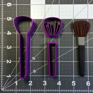 May include: Three purple plastic cookie cutters shaped like makeup brushes. The cutters are on a gray grid background.