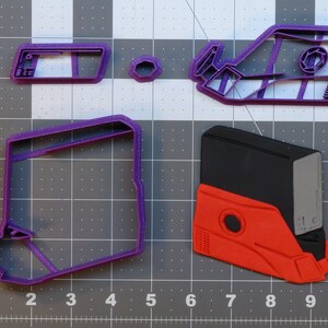 May include: A set of four purple plastic cookie cutters shaped like a spaceship, a rectangular box, a small circle, and a rectangular panel. The cookie cutters are on a grey grid background.