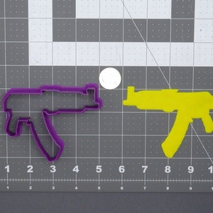 Puede incluir: Un cortador de galletas de metal morado con forma de pistola está junto a un recorte de fieltro amarillo con forma de pistola. Ambos están sobre una esterilla de corte gris con una regla. También hay una moneda.