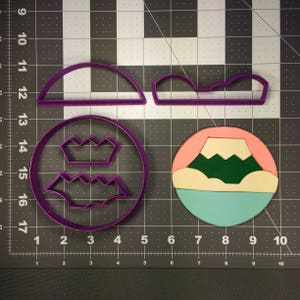 May include: Purple plastic cookie cutters in the shape of a mountain, a crown, and a wavy line. The cutters are on a gray grid background.