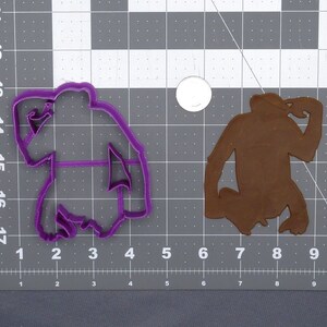 May include: Purple plastic cookie cutter shaped like a monkey sitting down with its hand on its head. The monkey is facing the left side of the image.