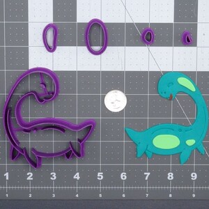 Puede incluir: Cortadores de galletas de plástico morado y verde con forma de plesiosaurios. Los cortadores están sobre una estera de corte gris con líneas de cuadrícula blancas.