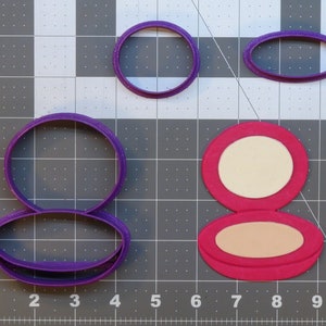 May include: Purple plastic cookie cutters in the shape of a round compact and a circle. A pink plastic cookie cutter in the shape of a round compact with a lid open.