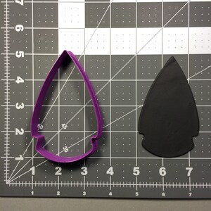May include: Purple plastic cookie cutter shaped like an arrowhead. The cutter is on a gray grid surface. A black arrowhead is to the right of the cutter.