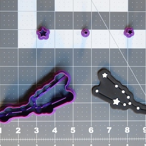 May include: A collection of purple plastic cookie cutters in various shapes, including a star, a circle, and a constellation design. The constellation cutter is black with white star accents. The items are arranged on a gray grid surface.