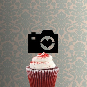 May include: A red velvet cupcake topped with white frosting and red sprinkles. A black camera with a heart in the lens is on top of the cupcake.