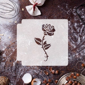 May include: White plastic stencil with a single rose design. The stencil is on a brown surface with flour scattered around it.
