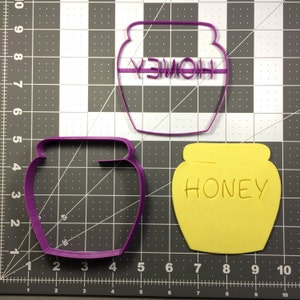 May include: Two cookie cutters in the shape of honey jars. The top cutter is purple and says "HONEY" in a stylized font. The bottom cutter is yellow and says "HONEY" in a simple font.