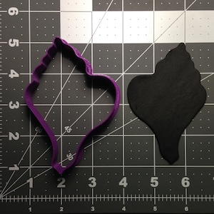 Puede incluir: Un cortador de galletas de plástico morado en forma de corazón estilizado con un borde superior festoneado, junto a un trozo de material negro en forma de corazón. El cortador está sobre una superficie de cuadrícula negra con medidas en centímetros.