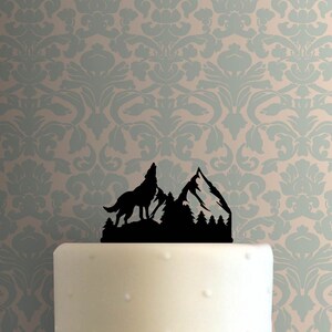 May include: Black silhouette cake topper of a wolf howling at the moon with mountains and trees in the background.