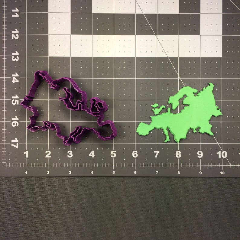 May include: A purple cookie cutter in the shape of a map of Europe is next to a green map of Europe. The cookie cutter is made of plastic and is on a black and gray grid surface. The green map is also made of plastic.