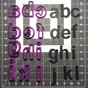 Pode incluir: Dois conjuntos de cortadores de biscoitos em forma de alfabeto, um roxo e outro preto. O conjunto roxo tem uma fonte mais estilizada, enquanto o conjunto preto tem uma fonte mais tradicional.
