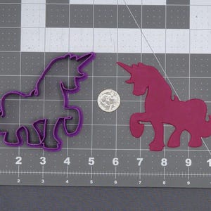 May include: A purple unicorn cookie cutter and a magenta unicorn cutout are displayed on a gray grid background. A coin is next to the unicorn cutout. The cookie cutter is approximately 13 cm long.