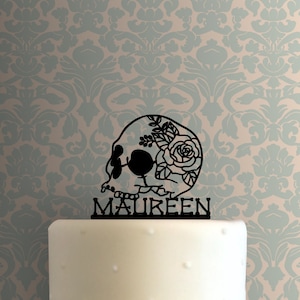 May include: Black skull cake topper with a floral design and the name "MAUREEN" written below.